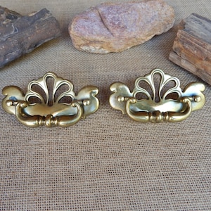 May include: Two antique brass drawer pulls with a decorative fan-shaped design. The pulls have a curved handle and are mounted on a decorative backplate. The hardware is a warm gold color and is set against a neutral background.