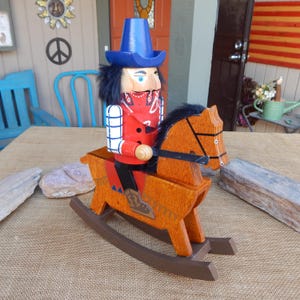 May include: A wooden cowboy nutcracker riding a rocking horse. The cowboy is wearing a blue hat, red bandana, and plaid shirt. The horse is brown with black accents.