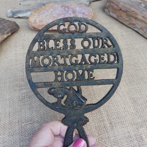 May include: A cast iron trivet with the words "GOD BLESS OUR MORTGAGED HOME" in a circular design. The trivet has a handle and a small house graphic. The metal has a dark, aged appearance.