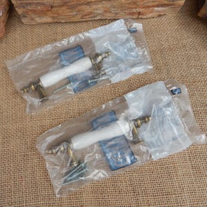 May include: Two cabinet pulls in clear plastic packaging. Each pull has a white ceramic handle and ornate brass-colored metal hardware. Screws and blue plastic pieces are also visible. The background is a textured tan surface.