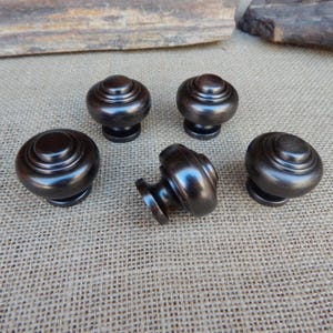 May include: Five bronze-colored cabinet knobs with a mushroom-shaped design. The knobs have a rounded top with concentric rings and a short, cylindrical base. One knob is turned on its side, showing the stem.