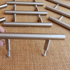 Op de afbeelding: Een set van tien kastgrepen van geborsteld nikkel, elk met een rechthoekige balk en een T-vormige basis. De grepen zijn in een verspringend patroon op een bruine jute ondergrond gerangschikt.