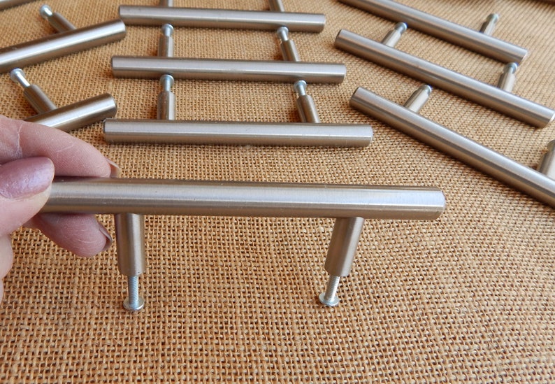 Op de afbeelding: Een set van tien kastgrepen van geborsteld nikkel, elk met een rechthoekige balk en een T-vormige basis. De grepen zijn in een verspringend patroon op een bruine jute ondergrond gerangschikt.