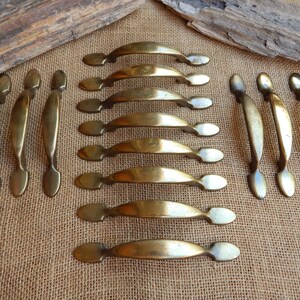 May include: A set of 14 brass cabinet pulls with a simple, elegant design. The pulls have a slightly curved shape and rounded ends. They are a classic style that would complement a variety of home decor.