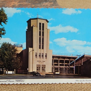 May include: A vintage postcard featuring a photo of the Memorial Tower at New Mexico State University (NMSU). The tower is a tall, rectangular building with a clock tower on top. The building is made of light-colored stone and has a large number of windows. The tower is surrounded by a large, open-air stadium with bleachers. The sky is blue with white clouds. The postcard is titled "Memorial Tower, NMSU" and is credited to "Photo by Lou Candelaria."