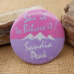 May include: A pink and purple round button with the text "Ski it to Believe it!" and "Sandia Peak Albuquerque, NM". The button features a graphic of three white mountains.