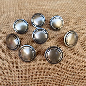 May include: A collection of nine antique brass-colored cabinet knobs. Each knob has a rounded, slightly domed center surrounded by a raised, circular border. The knobs are arranged on a textured, tan fabric surface.