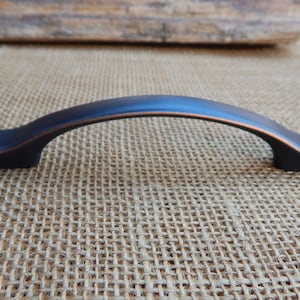 May include: A black and copper cabinet pull with a curved shape. The pull is made of metal and has a smooth finish.