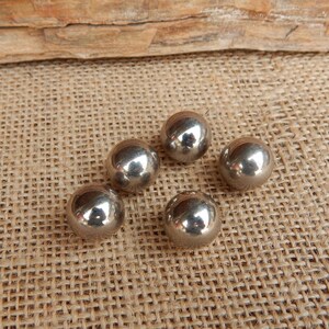 May include: Five shiny, silver metal spheres are scattered on a textured, tan surface. The spheres reflect light, creating highlights. The background features a piece of wood.