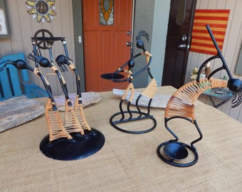 Peut inclure: Quatre bougeoirs en métal avec des figurines en paille tressée. Les figurines sont dans différentes poses, dont une femme tenant une assiette, une femme les bras levés et deux femmes dansant. Les bougeoirs sont noirs et ont un aspect rustique.