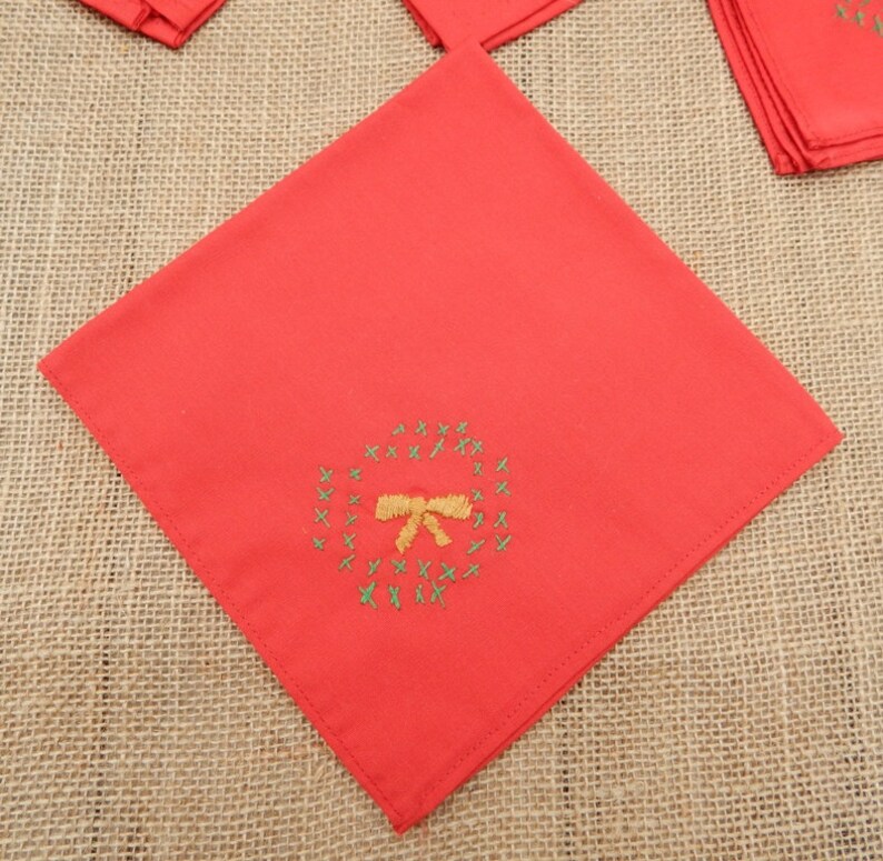 May include: Red cloth napkin with an embroidered wreath and bow design. The wreath is green, and the bow is gold. Several napkins are visible, suggesting a set. The napkins are square and made of a solid red fabric.