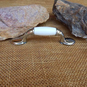 May include: A cabinet pull with a white ceramic center and polished chrome finish. The handle has a curved design and is suitable for drawers or cabinets. The hardware is shown on a textured, neutral background.