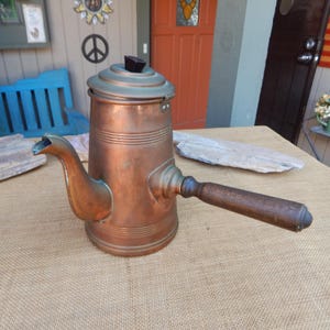 May include: A vintage copper coffee pot with a wooden handle. The pot has a rounded body with a spout and a lid. The handle is attached to the side of the pot.