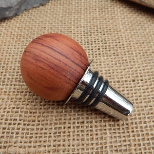 May include: A wooden bottle stopper with a round, reddish-brown wood top and a silver metal base with a black rubber ring.