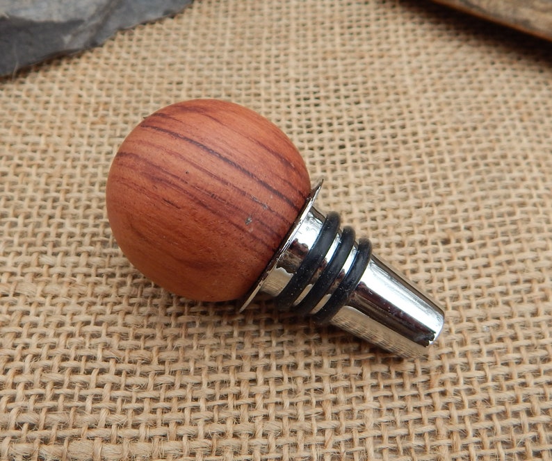 May include: A wooden bottle stopper with a round, reddish-brown wood top and a silver metal base with a black rubber ring.