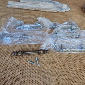 May include: Several antique brass cabinet pulls, some still in plastic packaging, are arranged on a textured, tan surface. The pulls have a detailed, ornate design. Two silver screws are also visible, suggesting they are ready for installation.