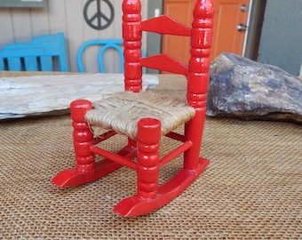 Peut inclure: Petite chaise à bascule en bois rouge avec une assise tressée. La chaise présente un design classique avec des montants et des bascules arrondis. La chaise est peinte en rouge vif et est probablement un objet décoratif.