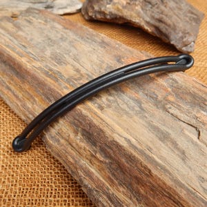 May include: A black hair clip with a curved, double-band design. The clip is made of a smooth, matte material and is resting on a piece of weathered wood. The clip's ends have rounded, bulbous shapes.