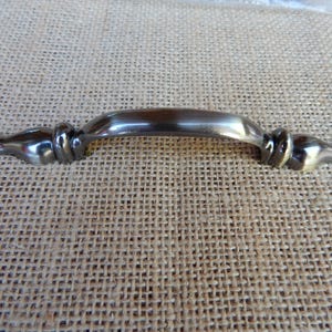 May include: A metallic drawer pull handle with a dark, brushed finish. The handle features a smooth, arched center section flanked by decorative, rounded accents on both ends, creating a balanced and symmetrical design for furniture hardware.