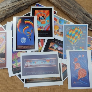 May include: A collection of colorful postcards featuring hot air balloons. The postcards are arranged in a pile, with some overlapping. The postcards are all different designs, but they all feature hot air balloons in various colors and patterns. Some of the postcards have text on them, including the words "Albuquerque International Balloon Fiesta".