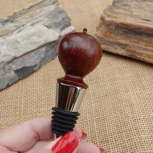 K&ouml;nnte beinhalten: Ein Flaschenverschluss aus Holz mit dunkelroter Oberfl&auml;che und einem silbernen Metallkragen. Der Verschluss hat eine abgerundete Oberseite und eine schwarze Gummibasis.