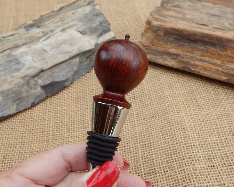 K&ouml;nnte beinhalten: Ein Flaschenverschluss aus Holz mit dunkelroter Oberfl&auml;che und einem silbernen Metallkragen. Der Verschluss hat eine abgerundete Oberseite und eine schwarze Gummibasis.