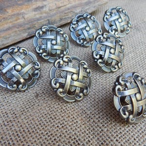 May include: A set of seven round metal buttons featuring an intricate, raised, interwoven lattice design with a metallic silver finish. Each button has a central circular stud surrounded by curved, braided bands and a textured outer border.