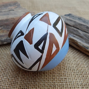 May include: A small, round ceramic pot with a white base and a geometric design in black, brown, and white. The pot has a light blue glaze on the bottom.