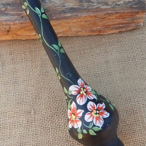 May include: Black ceramic vase with a long, slender neck and a bulbous base. The vase is hand-painted with white and red flowers and green leaves. The vase is signed on the bottom with the Hebrew word "O'shev".