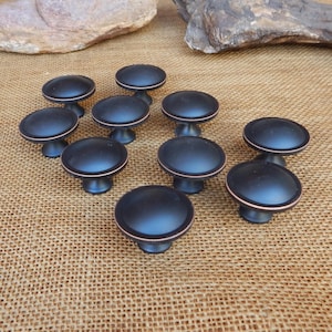 May include: A collection of ten black cabinet knobs with a copper-colored trim. The knobs have a rounded, mushroom-like shape and are arranged on a textured, tan surface. The hardware is likely for furniture or cabinetry.