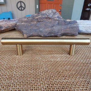 May include: A gold-colored, cylindrical cabinet pull with two short, cylindrical posts. The hardware is made of metal and is designed for use on drawers or cabinets. The pull is shown against a neutral background.