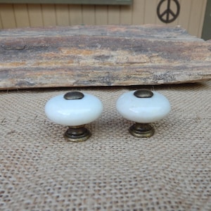 May include: Two round cabinet knobs featuring smooth, glossy white ceramic surfaces. Each knob is accented with a dark, antiqued brass-toned metal base and a matching circular metal cap at the center, providing a contrast to the white finish.