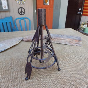 May include: A rustic, brown metal candle holder with a central spike and decorative cutouts. The holder features arrow-shaped accents and a circular base with a stylized design. The candle holder is designed to hold a single candle.