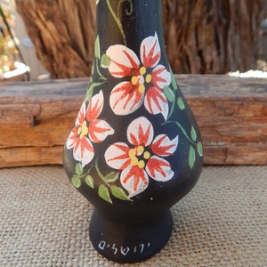 May include: Black ceramic vase with hand-painted red, white, and green flowers. The vase has a rounded body and a narrow neck. The bottom of the vase is signed in Hebrew.