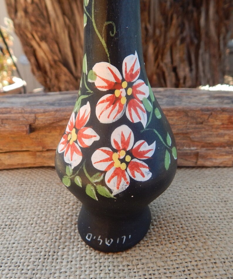 May include: Black ceramic vase with hand-painted red, white, and green flowers. The vase has a rounded body and a narrow neck. The bottom of the vase is signed in Hebrew.
