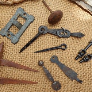 May include: A collection of antique metal hardware pieces, including a door latch, a hook, a pair of hinges, a door handle, and a small nail. The pieces are arranged on a burlap background.