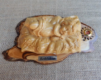 Könnte beinhalten: Eine geschnitzte Holz-Weihnachtskrippe mit einem Baby in einer Krippe. Die Skulptur ist hellbraun und ruht auf einem Holzsockel mit dem Wort "Bethlehem" auf einem kleinen Metallschild. Ein dekoratives Element mit goldenem Zentrum befindet sich auf der rechten Seite.