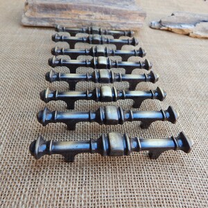 May include: A row of ten antique brass cabinet pulls with a decorative design. The pulls are arranged in a line on a burlap surface.