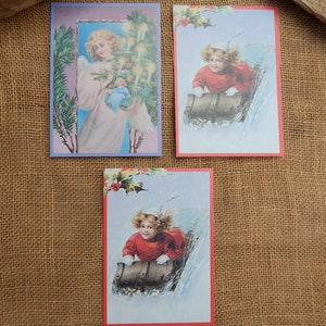 May include: Three vintage Christmas postcards. One features an angel holding a doll in front of a Christmas tree. The other two feature children sledding down a snowy hill.