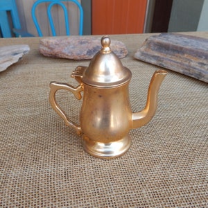May include: A small gold-colored teapot with a hinged lid and a spout. The teapot is sitting on a brown burlap surface.
