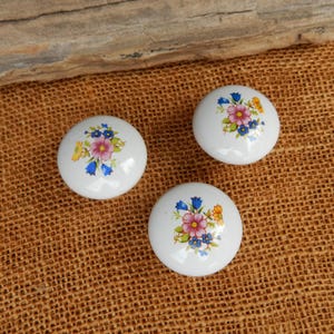 May include: Three white ceramic cabinet knobs with a floral design featuring pink, blue, and yellow flowers. The knobs are round and have a glossy finish, perfect for adding a touch of elegance to furniture.
