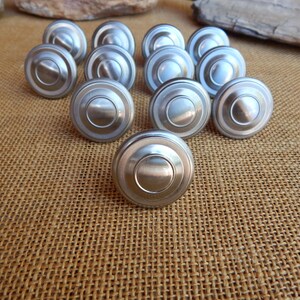 May include: A set of 12 silver-toned round cabinet knobs with a raised outer ring and a smooth, slightly domed center. The knobs are arranged in a semi-circle on a brown burlap surface.
