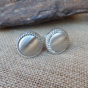 May include: Two silver-toned cufflinks with a circular design. Each cufflink features a smooth, brushed metal center surrounded by a braided rope-like border. The cufflinks are displayed on a textured, tan surface.