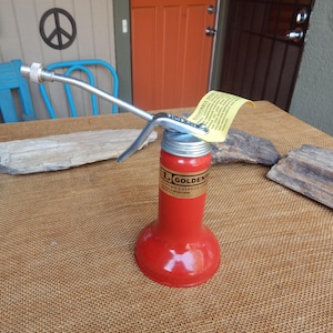 May include: A vintage red oil can with a silver metal spout and handle. The can has a label that reads "GOLDEN" and "BUTTON-LAINSON CO." A yellow tag is attached to the top. The can is sitting on a brown surface.