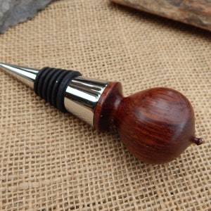 K&ouml;nnte beinhalten: Ein Flaschenverschluss aus Holz mit dunkelroter Oberfl&auml;che und einem silbernen Metalldeckel. Der Verschluss hat eine Gummidichtung, um den Wein frisch zu halten.