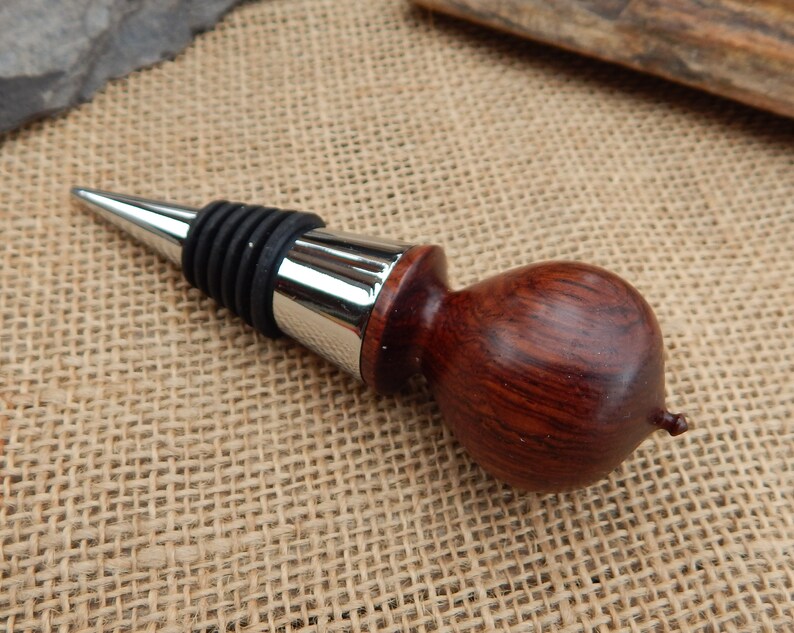 K&ouml;nnte beinhalten: Ein Flaschenverschluss aus Holz mit dunkelroter Oberfl&auml;che und einem silbernen Metalldeckel. Der Verschluss hat eine Gummidichtung, um den Wein frisch zu halten.