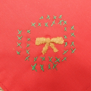 May include: A red fabric square with an embroidered Christmas wreath design. The wreath is made of green cross-stitches, surrounding a gold bow. The fabric has a slightly textured appearance.