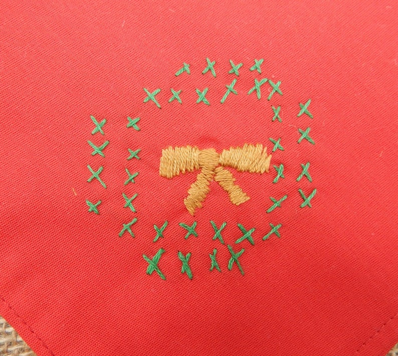 May include: A red fabric square with an embroidered Christmas wreath design. The wreath is made of green cross-stitches, surrounding a gold bow. The fabric has a slightly textured appearance.