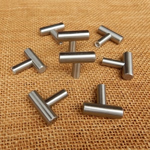 May include: A collection of brushed nickel T-bar cabinet pulls. The hardware is cylindrical with a T-shaped design. The pulls are scattered on a textured, tan surface.