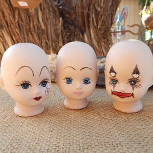 May include: Three porcelain doll heads with painted faces. The doll heads have blue eyes, painted eyebrows, and red lips. One doll head has black and white painted markings around the eyes.